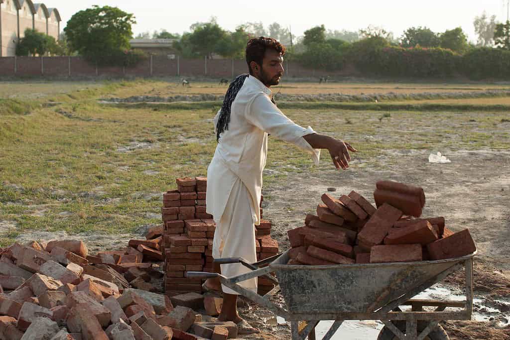 Worker loads wheel barrel full of bricks in rural Pakistani construction site.
