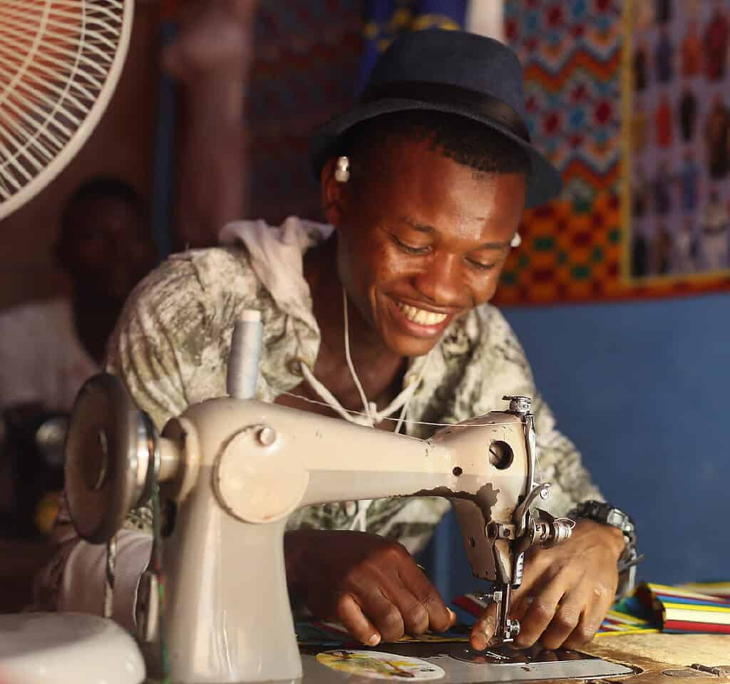 Man cheerfully sews clothes in West African garment shop while listening to music