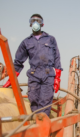 Man wearing respiration mask and other protective gear resting on top of farming equipment in a open field