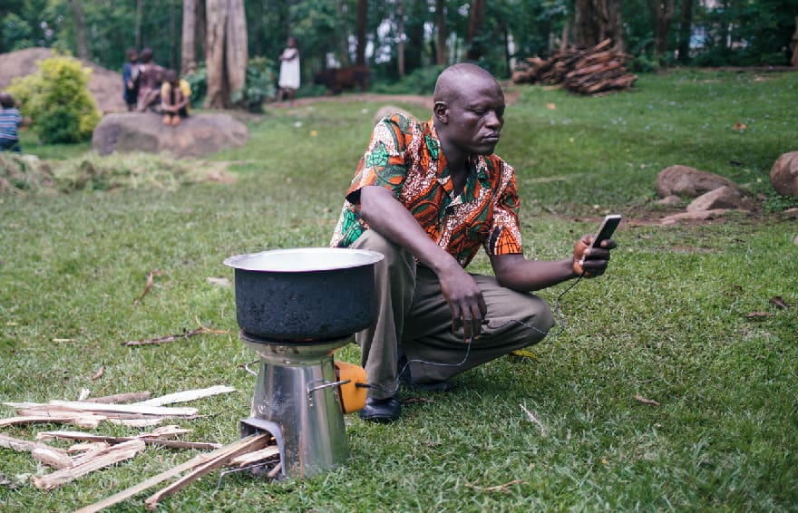 Kneeling man checks phone that is being charged by a cookstove that is also heating a pot