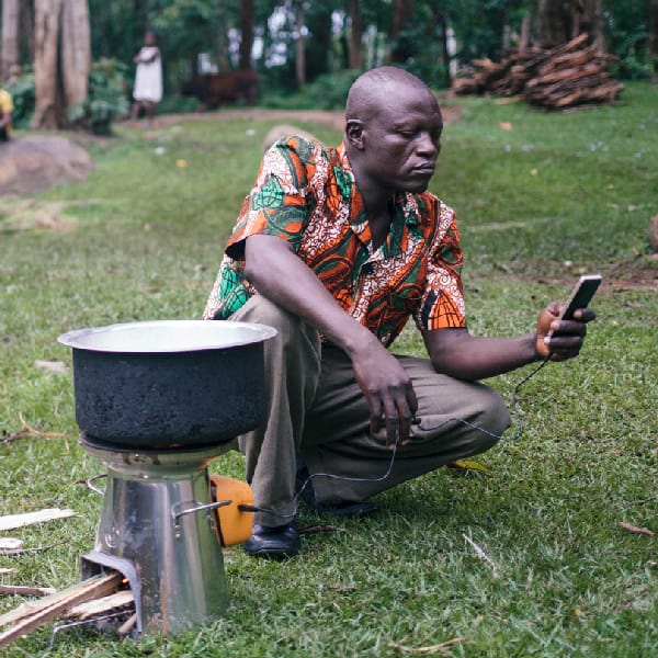 Kneeling man checks phone that is being charged by a cookstove that is also heating a pot