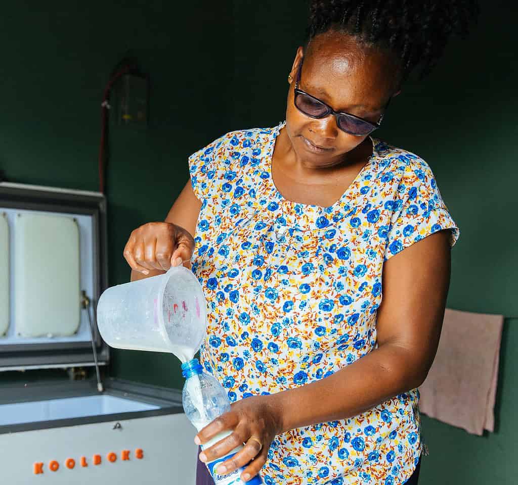 Woman carefully pours milk into a plastic bottle next to an open Koolboks refrigeration unit in her West Africa home