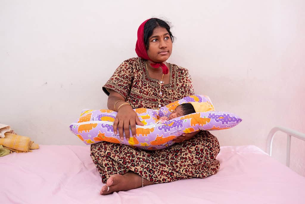 Women sits on bed with her child in a swaddle in Indian home
