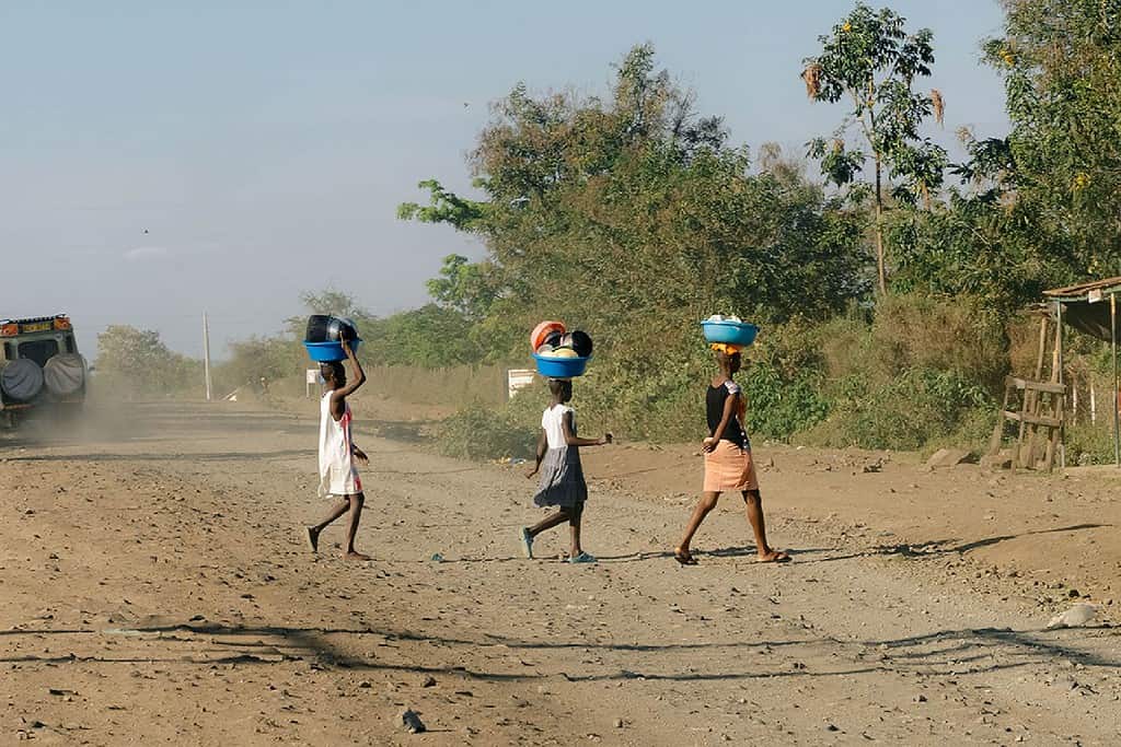 Three women cross a East African dirt road while carrying various bowls on top of their heads