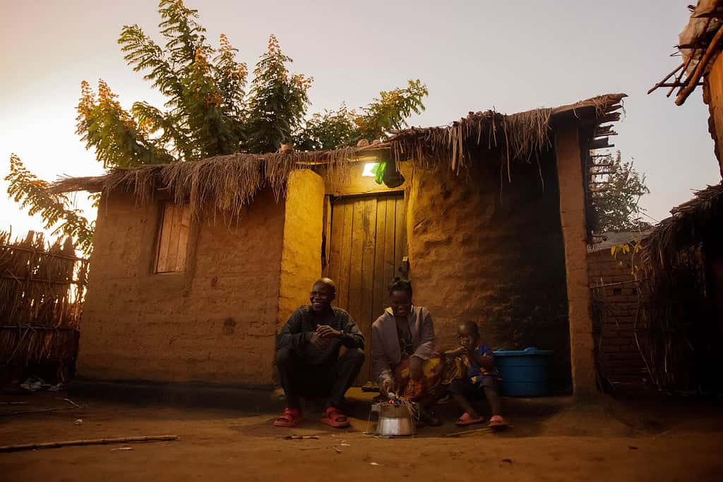 Family sits outside hut while starting cookstove under a solar light at dusk; Photo courtesy of Yellow