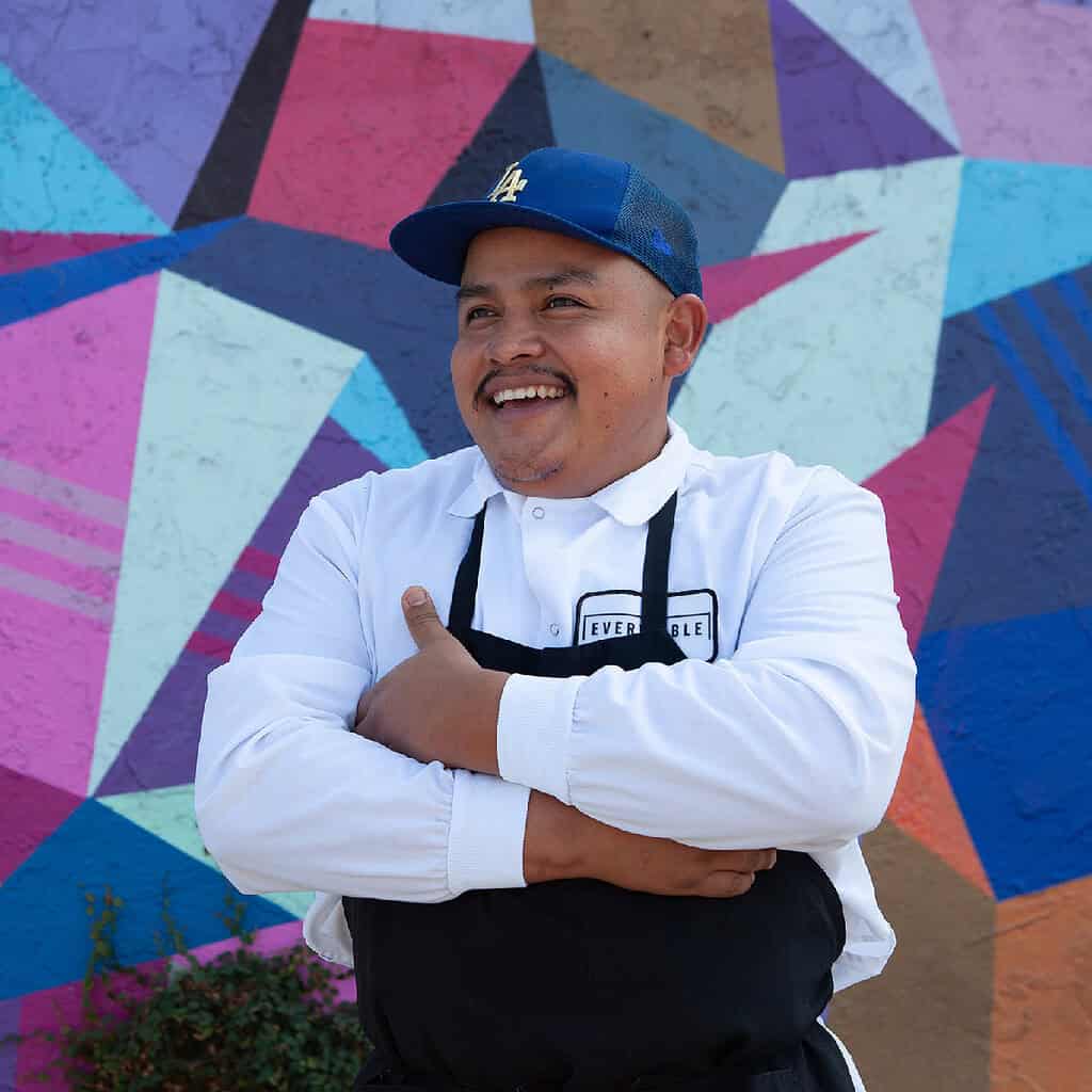 A man in a white shirt and apron stands in front of a colorful geometric mural, smiling with his arms crossed.