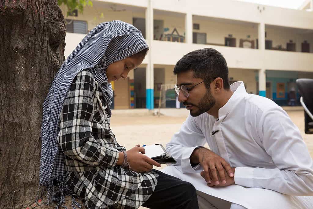 A child uses a tablet underneath a tree in a school yard, while a teacher looks on