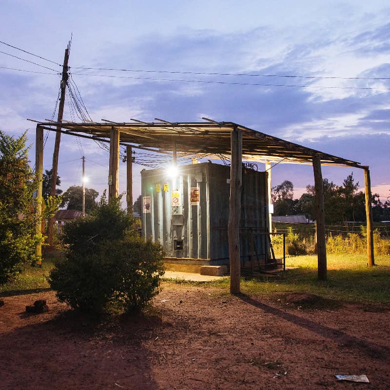 A small structure with a roof, illuminated at dusk, surrounded by trees and electric poles. Possible location: sub-Saharan Africa.