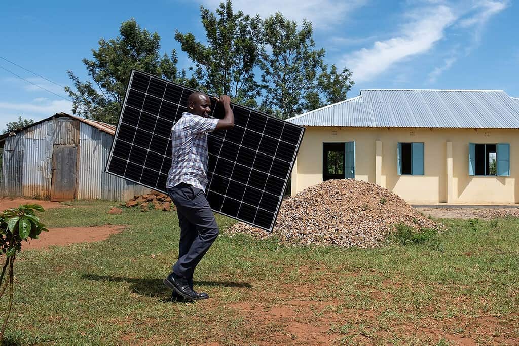 A person is carrying a solar panel in a rural setting, highlighting renewable energy efforts.