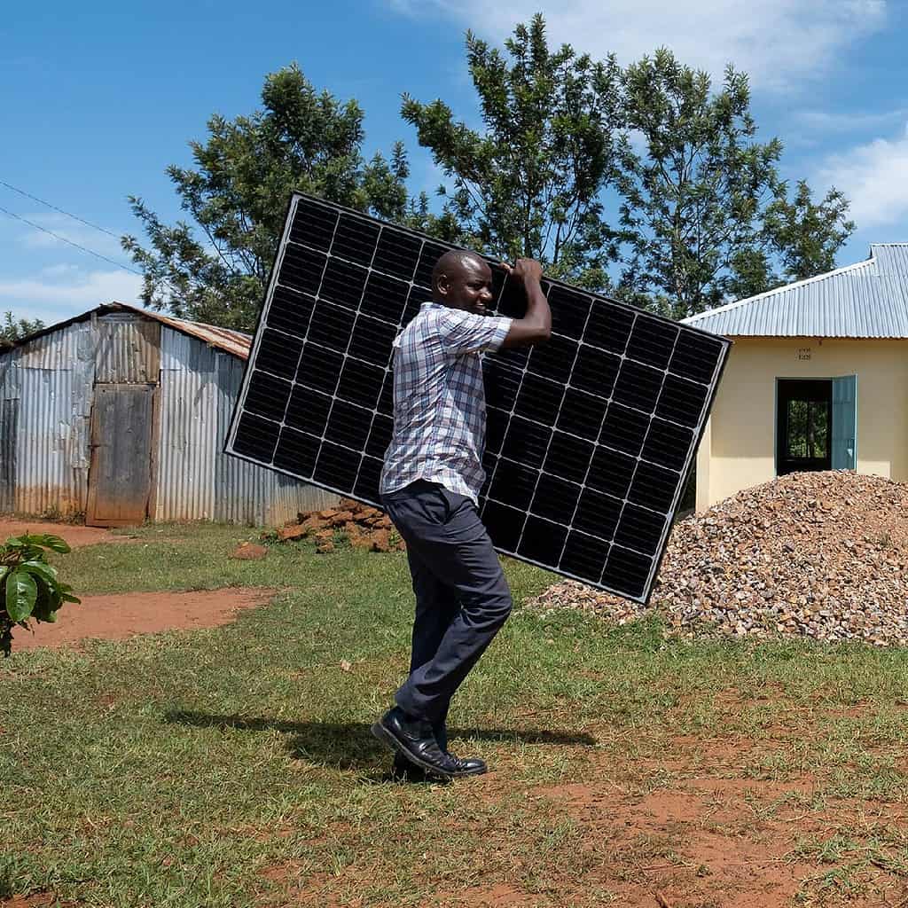 A person is carrying a solar panel in a rural setting, highlighting renewable energy efforts.