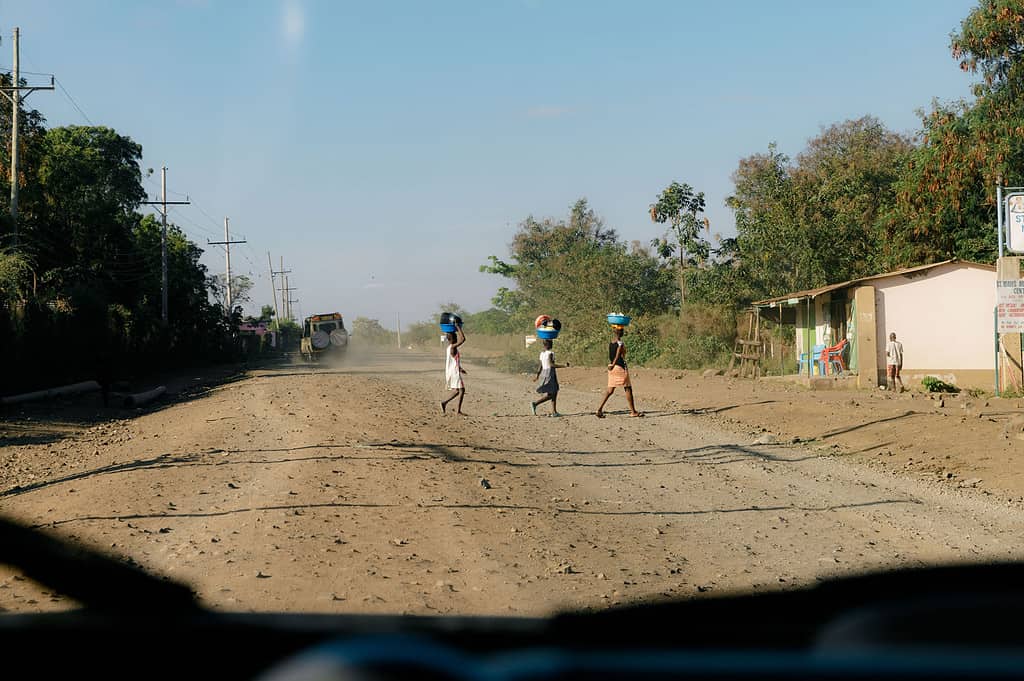 Three women cross a East African dirt road while carrying various bowls on top of their heads