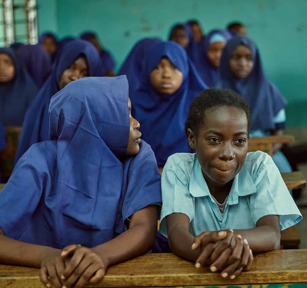 Students in a classroom, with some wearing blue hijabs, sitting at wooden desks.