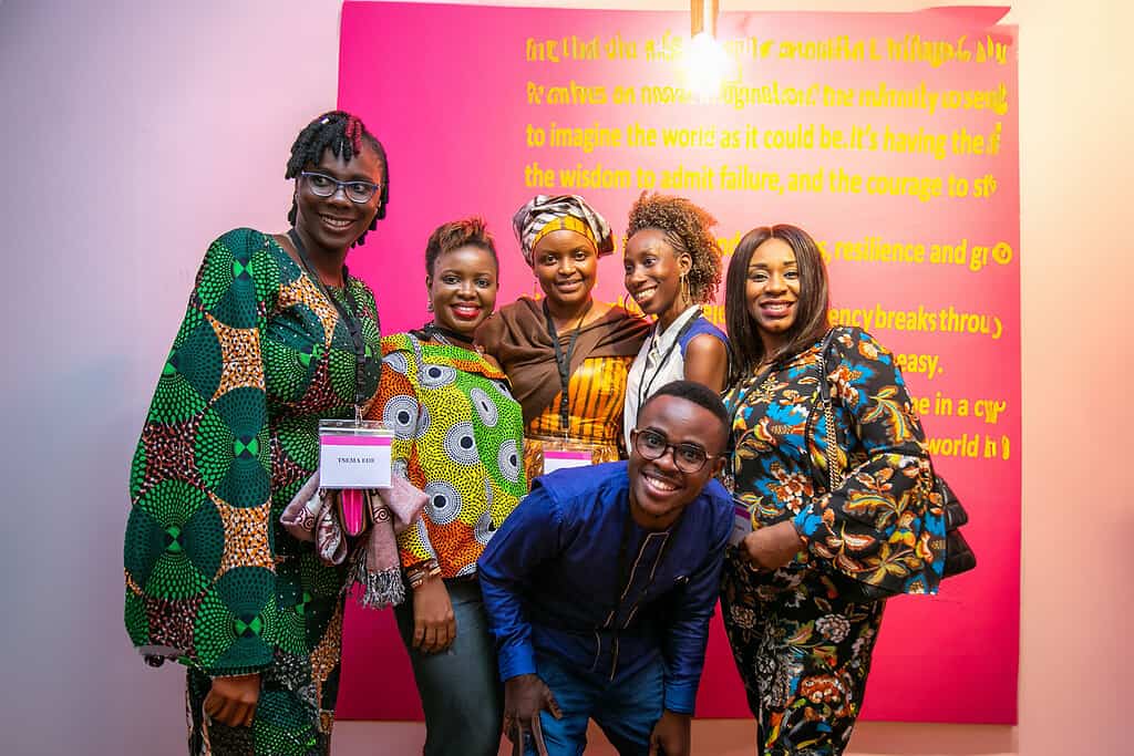 A group of people smiling and posing in colorful attire in front of a pink and yellow text display.