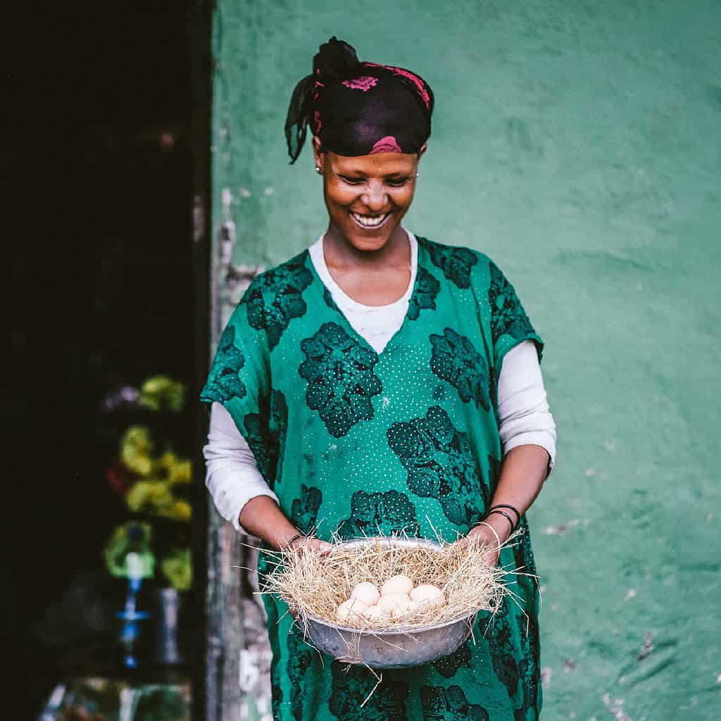 A smiling person holding a bowl with hay and eggs, standing against a green wall.