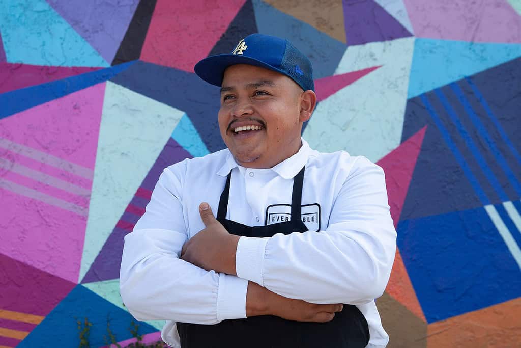 A man in a white shirt and apron stands in front of a colorful geometric mural, smiling with his arms crossed.