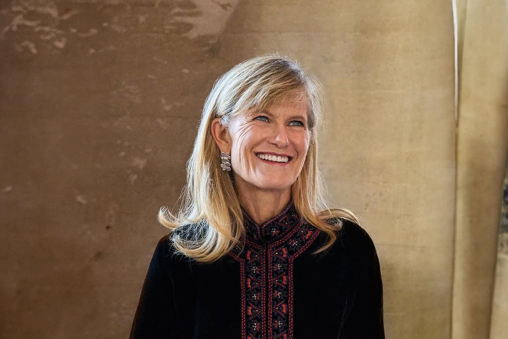 Jacqueline Novogratz smiling while looking away from the camera, wearing a dark patterned top.