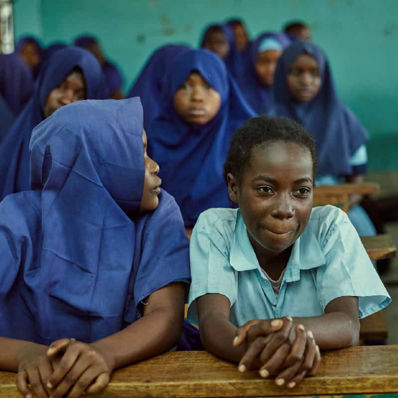 Students in a classroom, with some wearing blue hijabs, sitting at wooden desks.