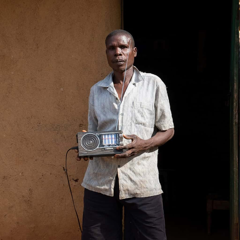 A person is holding a portable radio with an earphone in one ear, standing in front of a dark entrance.