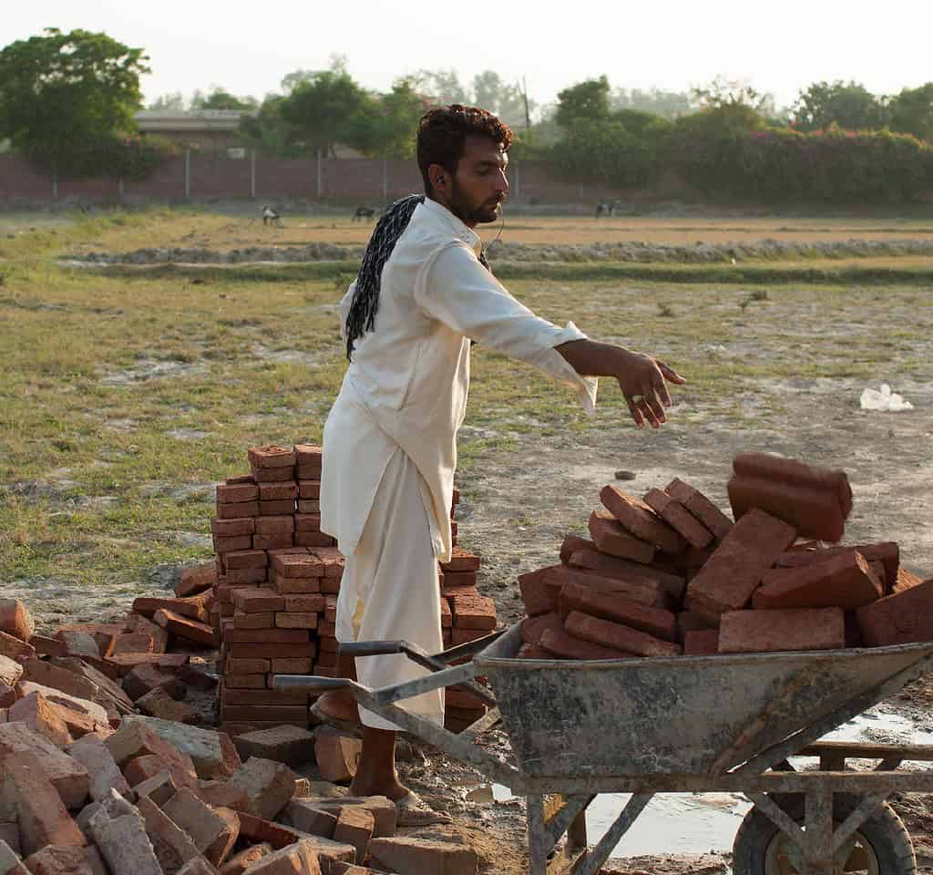 Worker loads wheel barrel full of bricks in rural Pakistani construction site.