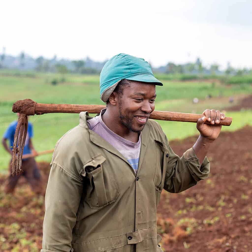 A smiling farmer carries a hoe over his shoulder in a green field.