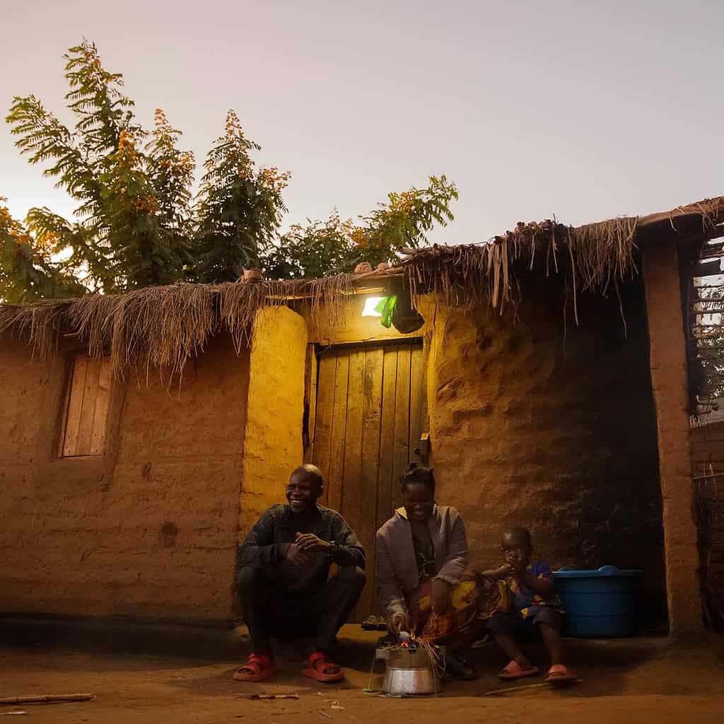 A family is sitting outside a traditional mud hut in the evening, with a lit bulb over the entrance. Sub-Saharan Africa context.