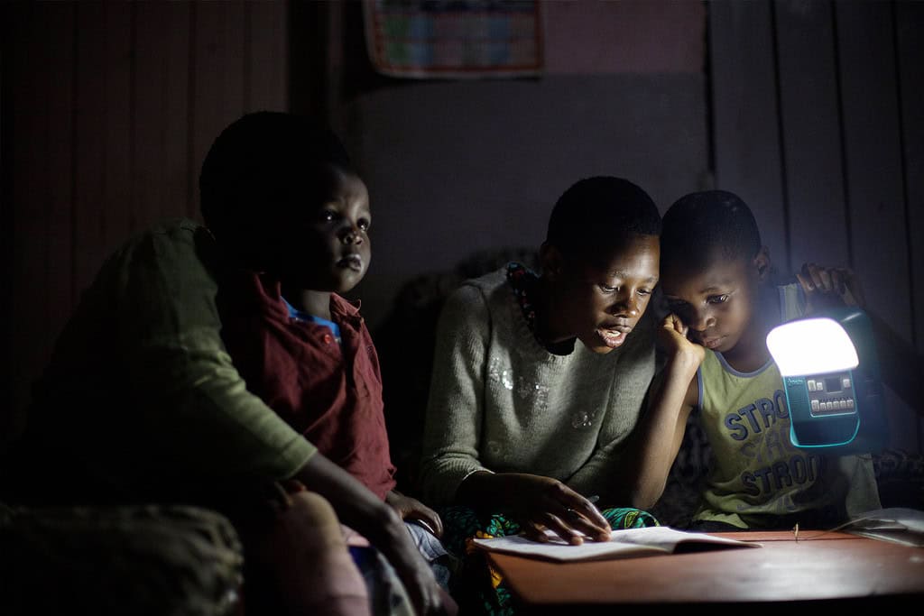Three children are reading a book together by the light of a lamp, possibly in a setting related to Sub-Saharan Africa.