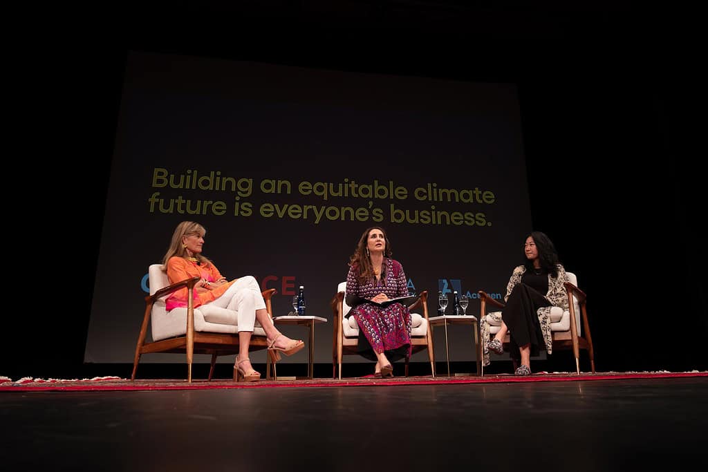 Three women, including Jacqueline Novogratz, are seated on a stage discussing climate equity, with the backdrop message promoting an equitable climate future.