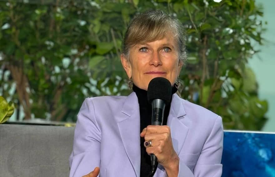 Jacqueline Novogratz holding a microphone, sitting in front of greenery, wearing a light purple blazer.