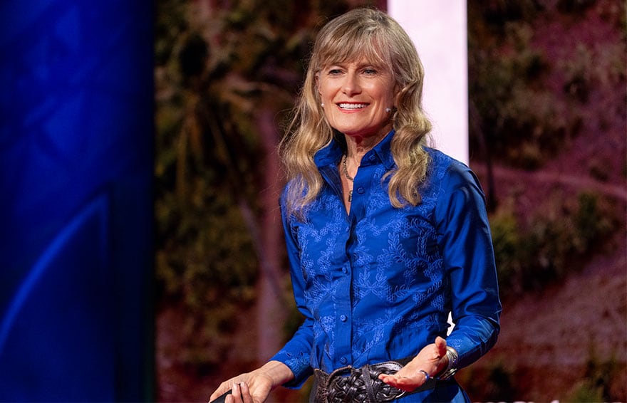Jacqueline Novogratz in a blue shirt is speaking on stage with a microphone headset.