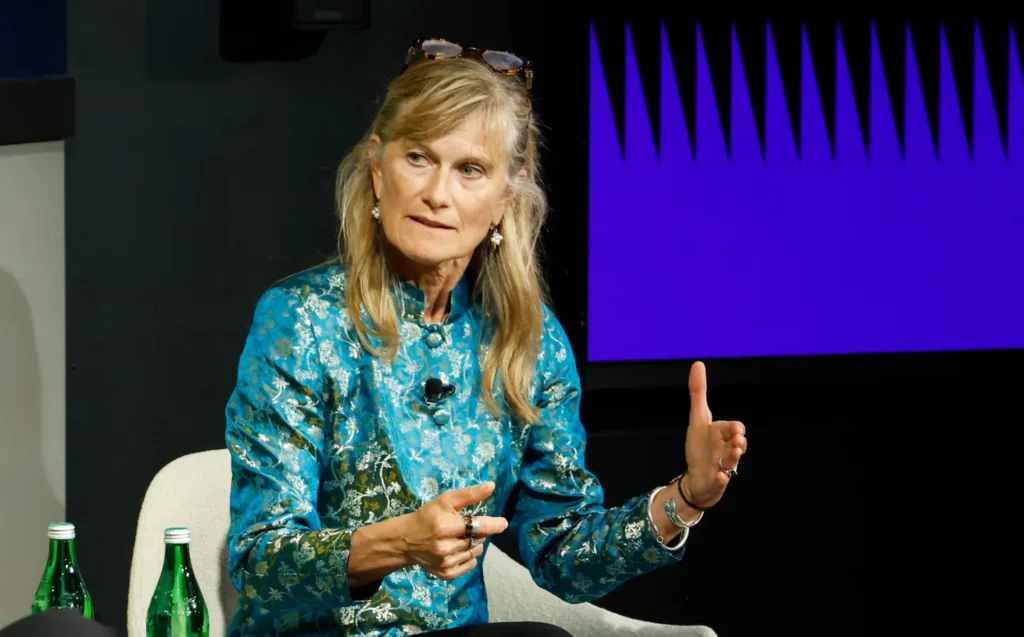 Jacqueline Novogratz in a blue jacket is speaking in a seated setting with two water bottles nearby.
